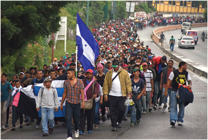 Hundreds of people walk on a roadway. In front, a Honduran flag and banner are carried by several people; others are carrying bags or luggage.