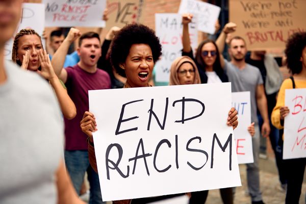 An African American woman is holding an end racism placard while protesting with a crowd of people on city streets.