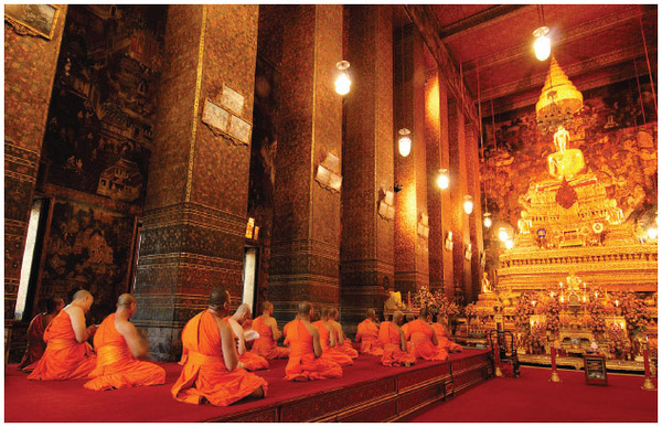 Buddhist monks are shown kneeling before an image of the Buddha in Wat Pho Temple in Bangkok, Thailand.